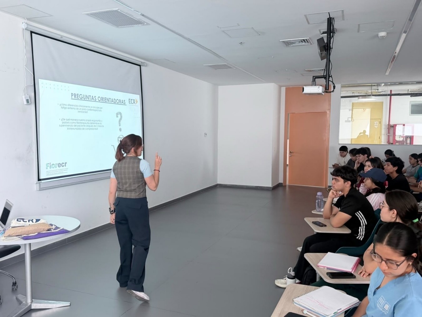 Profesora explica frente a una diapositiva titulada «Preguntas orientadoras» ante estudiantes sentados en un salón de clase